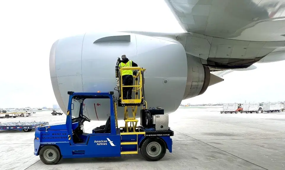 A worker in a high-visibility vest stands on a yellow lift attached to a blue American Airlines Harlan APV while performing maintenance on a massive jet engine.