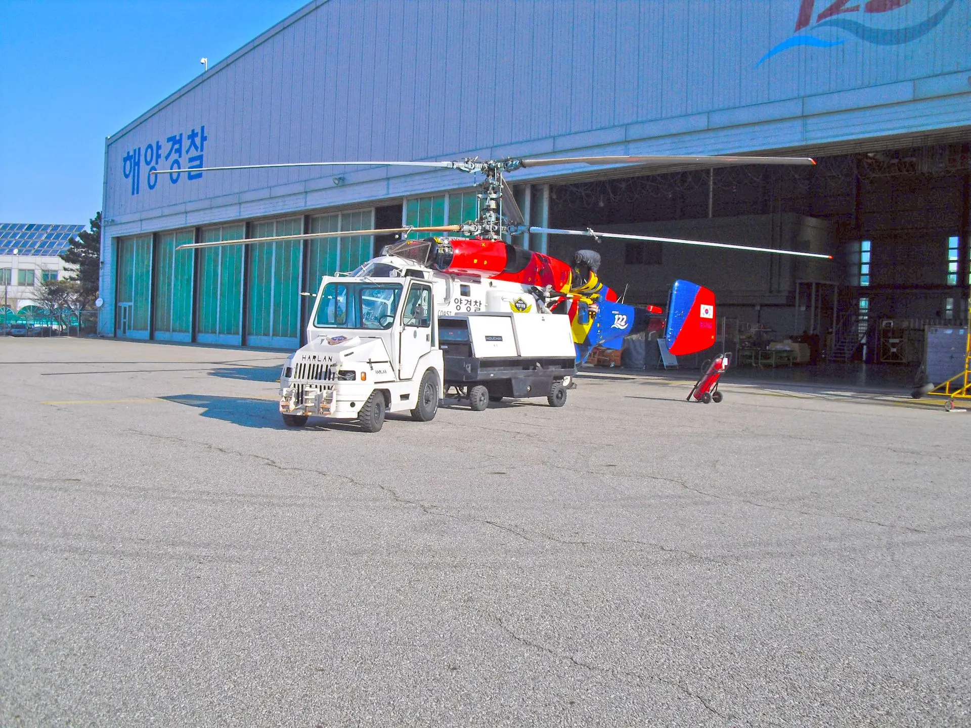 A white Harlan tractor is shown in the center of the frame, towing a brightly colored red, blue, and yellow helicopter across a sunny tarmac in front of a large hangar.