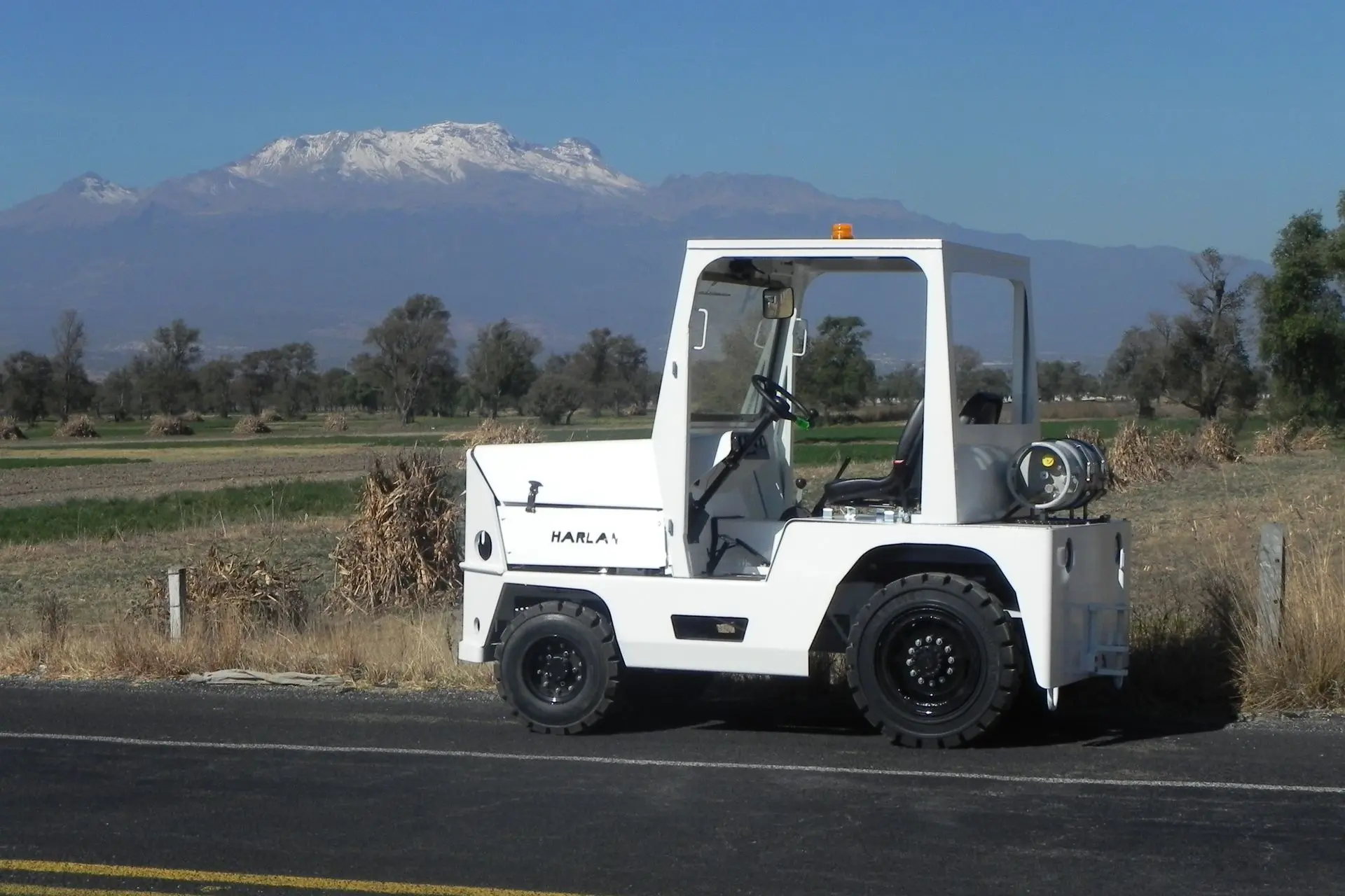 A white Harlan tractor with a black leather seat and a propane tank is parked on the edge of an asphalt road, with a scenic backdrop of fields and a snow-capped mountain.
