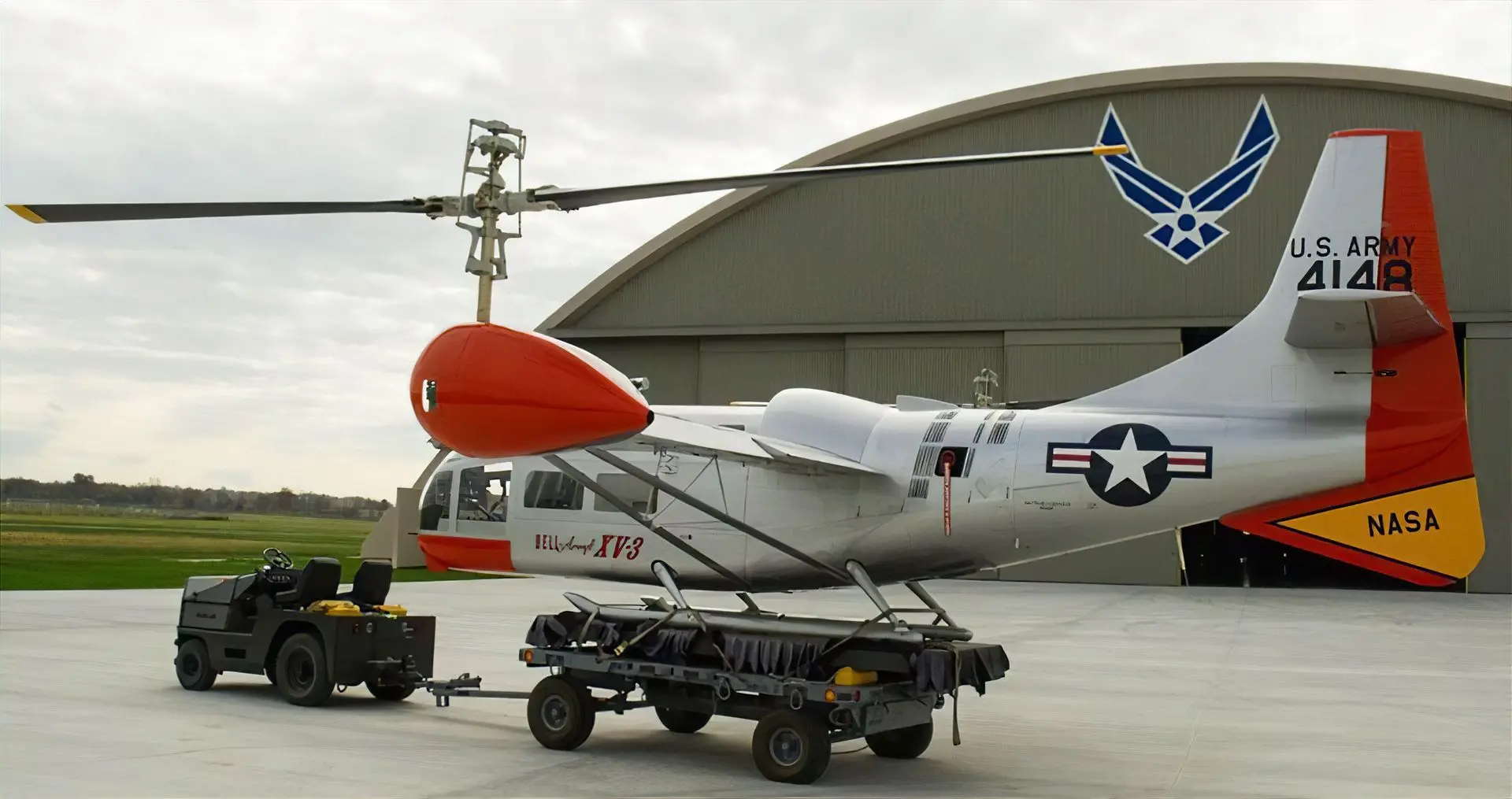 A dark grey Harlan tractor is shown in profile, towing a vintage Bell XV-3 aircraft on a trailer across an airfield toward a large hangar.