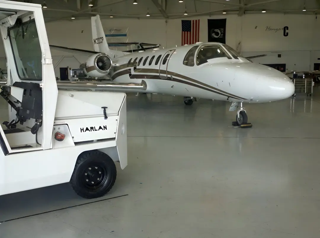 A white Harlan tractor is positioned in the foreground of a bright hangar, parked near a small white and tan private jet.