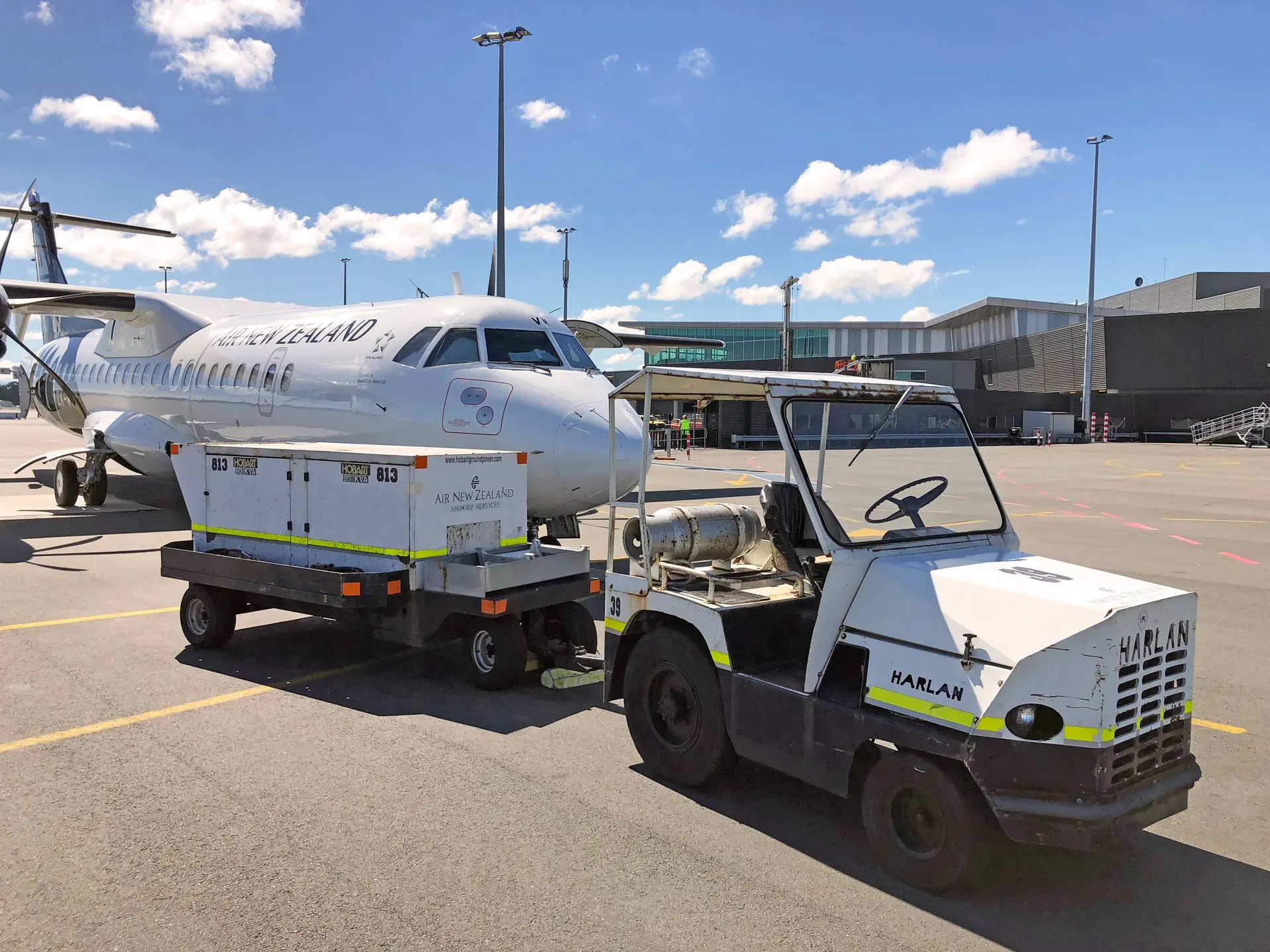 A white and black Harlan tractor with a propane tank is shown in the foreground, towing a large "Air New Zealand" power unit on a sunny tarmac in front of a white Air New Zealand passenger plane.