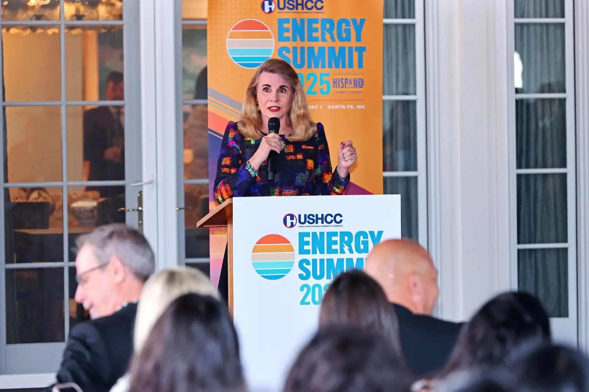 A woman with blonde hair and a colorful, textured blazer stands at a white podium, delivering a speech during the USHCC Energy Summit.