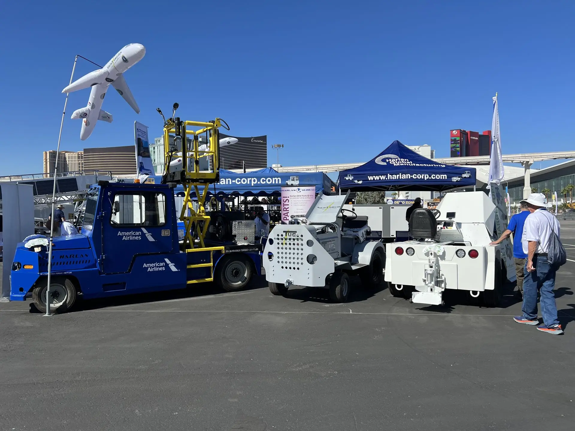 Various Harlan ground support vehicles are displayed at an outdoor expo under a clear blue sky, including a blue American Airlines tractor with a yellow scissor lift, a grey tug, and a white service vehicle.