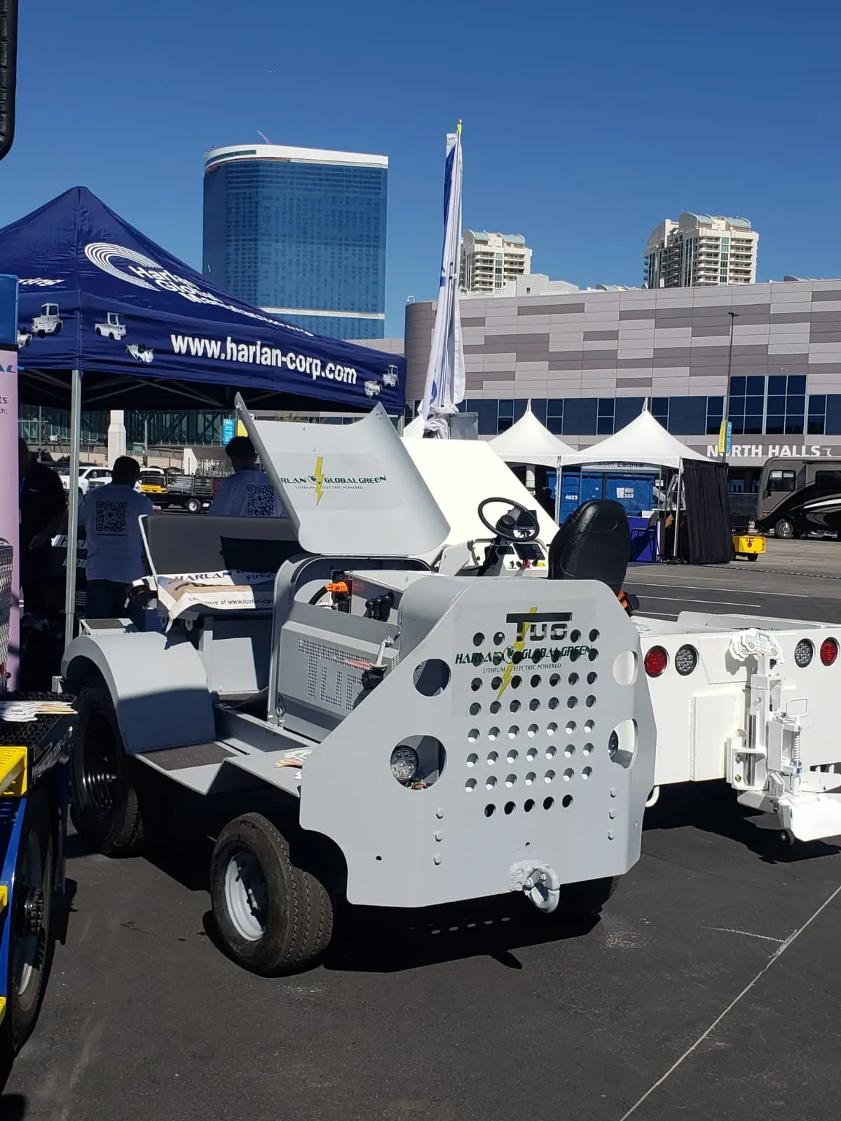A close-up photograph of a grey airport tractor refit with a Harlan Global Green E-conversion kit, displayed outdoors at a GSE Expo with its front panels removed to show the electric components.