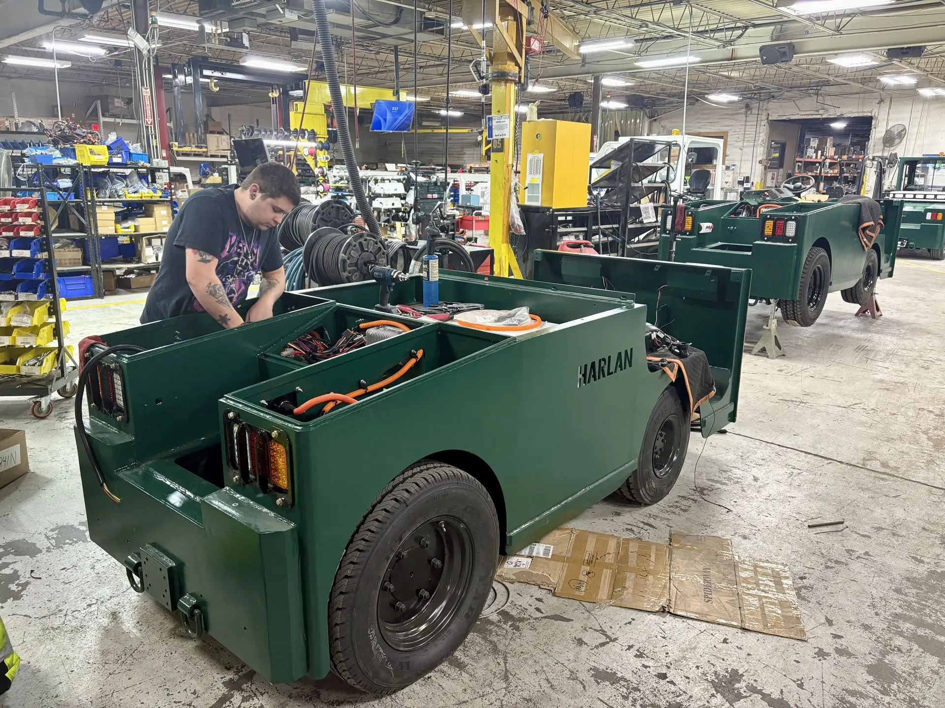 Assembly line technician installing electrical components into a fleet of green Harlan electric baggage tractors for airport ground support.