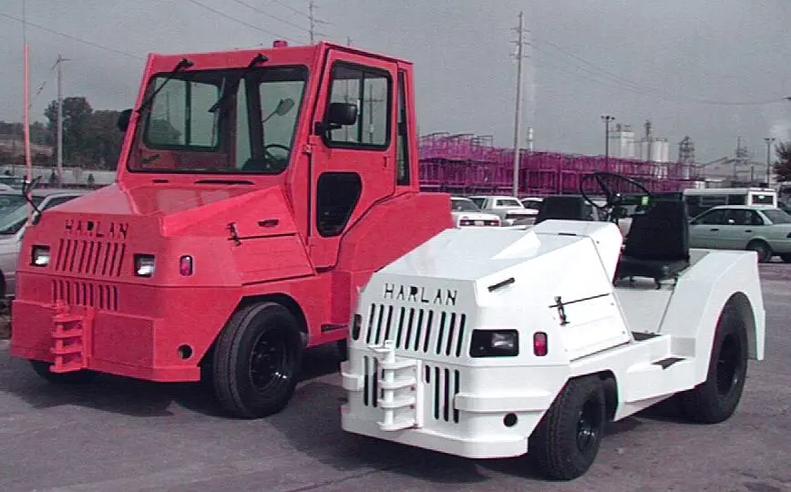 A red Harlan HLP tow tractor with an enclosed cab parked next to a compact white model, demonstrating the diverse scale and specialized configurations of heavy-duty airport towing vehicles.