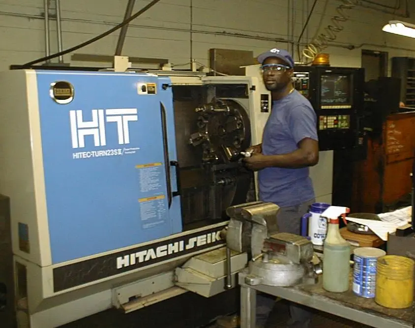 A plant technician operating a Hitachi Seiki Hitec-Turn 23SII CNC machine, demonstrating the rigorous manufacturing standards and technical skill behind Harlan industrial tractor production.